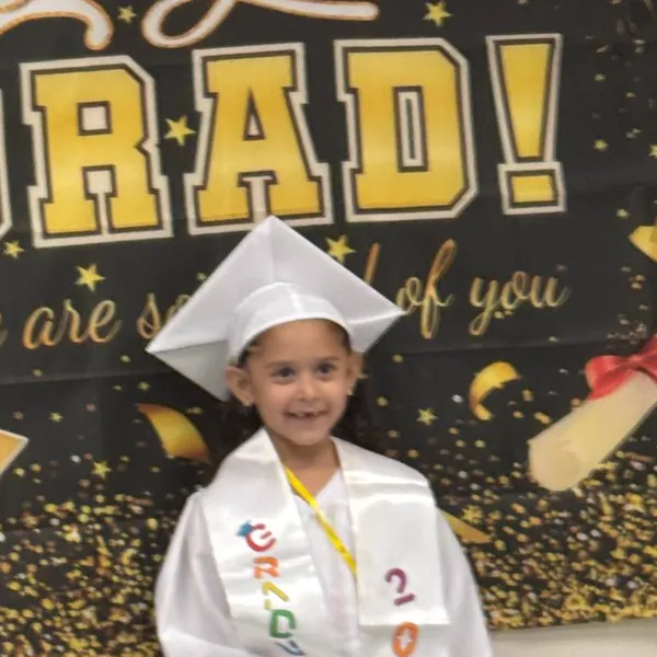 Smiling Rahmah preschool graduate in a white cap and gown